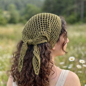 May include: Olive green crochet head covering with a net-like design. The head covering is tied at the nape of the neck with long, decorative ties. The background is a field of green grass and white flowers.