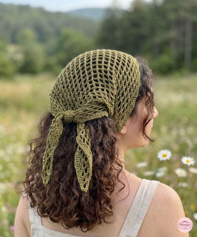 May include: Olive green crochet head covering with a net-like design. The head covering is tied at the nape of the neck with long, decorative ties. The background is a field of green grass and white flowers.