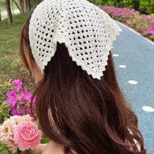 May include: A white, open-weave crochet headscarf. The head covering is triangular in shape and has a scalloped edge. The headscarf is worn on the head and covers the top of the head and the back of the hair. The headscarf is made of a soft, lightweight material.