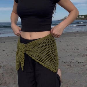 May include: Olive green crochet wrap tied at the waist, paired with black pants and a black crop top. The open weave of the wrap is visible. The background shows a beach and ocean.