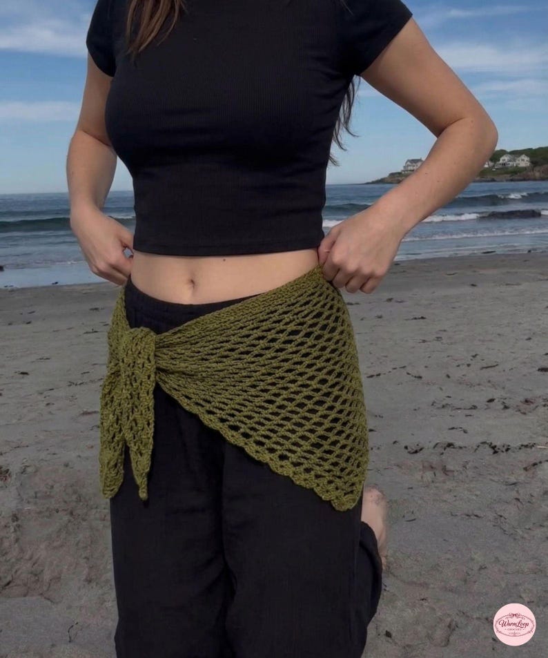 May include: Olive green crochet wrap tied at the waist, paired with black pants and a black crop top. The open weave of the wrap is visible. The background shows a beach and ocean.