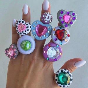 May include: A hand displaying a collection of colorful, chunky rings. The rings feature various designs, including hearts, polka dots, and a letter "D". Embellishments include rhinestones and large, colorful gems. The rings are in shades of pink, purple, green, and blue.