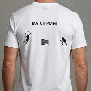 Puede incluir: Camiseta blanca con la frase "MATCH POINT" y gráficos negros con temática de tenis. El diseño incluye siluetas de jugadores de tenis y una red. La camiseta es de material suave.