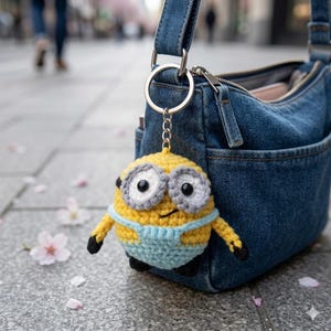 Könnte beinhalten: Ein gelber Minion-Schlüsselanhänger aus Häkelarbeit mit grauen und schwarzen Details, der an einer Jeans-Tasche hängt. Der Minion hat große Augen, eine blaue Latzhose und schwarze Füße. Der Schlüsselanhänger ist mit einem silbernen Ring und einer Kette befestigt.