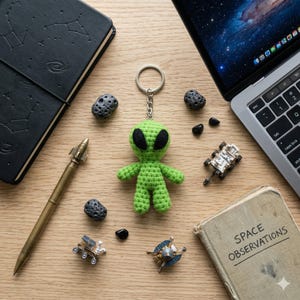 Könnte beinhalten: Ein grüner gehäkelter Alien-Schlüsselanhänger mit schwarzen Augen und einem silbernen Ring. Das Bild enthält auch ein Notizbuch, einen Stift, einen Laptop, kleine Weltraumfiguren und ein Vintage-Buch mit dem Titel "SPACE OBSERVATIONS" auf einer Holzoberfläche.