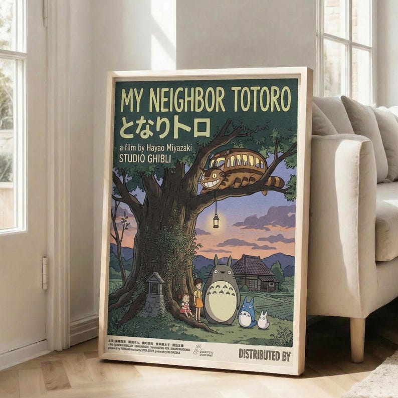 May include: Framed poster displaying beloved animated characters including large gray Totoro, smaller blue and white creatures, and two children standing beneath a large tree with the Cat Bus perched in branches. Artwork shows twilight countryside scene with traditional house, mountains, and warm sunset colors. Wooden frame leans against white wall next to beige couch in bright, modern living room with large windows.