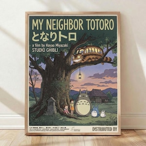May include: Framed Studio Ghibli poster print leaning against white wall on light wood floor. Features My Neighbor Totoro artwork with large tree, cat bus in branches with glowing windows, two children and Totoro characters beneath. Soft twilight colors in purple, green, and cream. Japanese text visible. Natural wood frame. Warm lighting creates gentle shadows on textured wall.