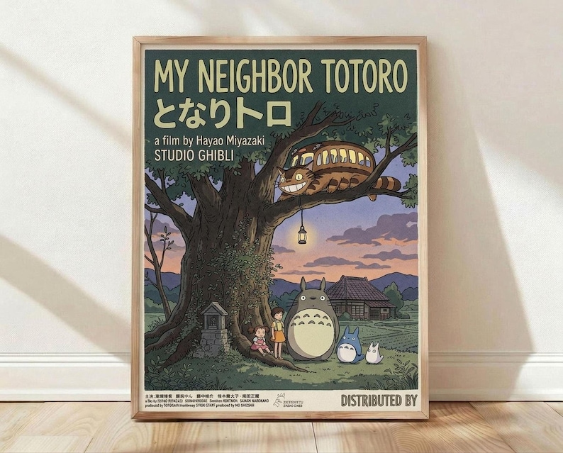 May include: Framed Studio Ghibli poster print leaning against white wall on light wood floor. Features My Neighbor Totoro artwork with large tree, cat bus in branches with glowing windows, two children and Totoro characters beneath. Soft twilight colors in purple, green, and cream. Japanese text visible. Natural wood frame. Warm lighting creates gentle shadows on textured wall.