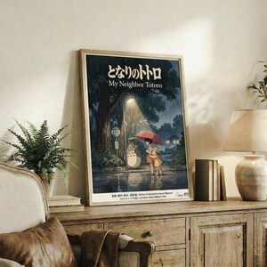 May include: Framed Studio Ghibli movie poster featuring Totoro and two children with red umbrella standing at rainy bus stop under large tree. Poster displays in light wood frame on rustic wooden console table beside cream armchair, fern plant, and ceramic vases in cozy living room setting with neutral walls.