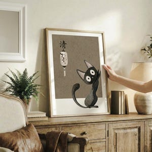 May include: Framed poster featuring a black cat with white eyes sitting beneath a decorative lantern and Japanese character on brown textured background, displayed on rustic wooden sideboard alongside fern plant, books, and ceramic vase in neutral-toned living room with beige furniture and walls