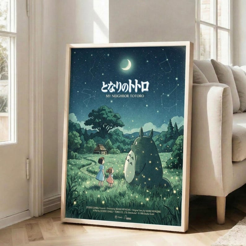 May include: A framed poster in a bright living room showing two children and Totoro in a moonlit forest scene with fireflies. The artwork features a starry night sky with crescent moon above rolling green hills, trees, and a small house. The large forest spirit character stands beside the children in the grass. Japanese text appears at the top. The wooden-framed poster leans against a white wall next to a beige couch near tall windows.