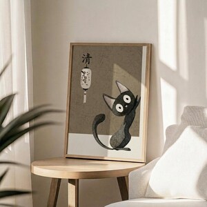 May include: Framed poster print featuring a stylized black cat with large white eyes and pointed ears sitting upright, displayed on a round wooden side table next to a beige upholstered chair. The artwork has a muted beige and gray color palette with Japanese characters at the top and a white paper lantern illustration. Natural sunlight streams across the minimalist room setting with white walls and a plant visible in the corner.