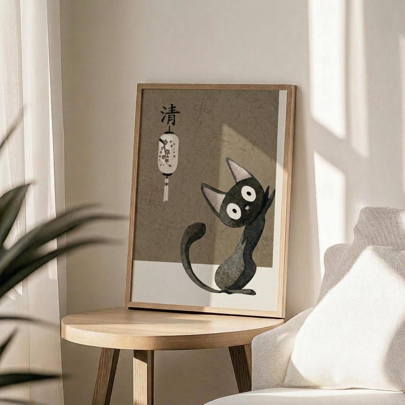 May include: Framed poster print featuring a stylized black cat with large white eyes and pointed ears sitting upright, displayed on a round wooden side table next to a beige upholstered chair. The artwork has a muted beige and gray color palette with Japanese characters at the top and a white paper lantern illustration. Natural sunlight streams across the minimalist room setting with white walls and a plant visible in the corner.
