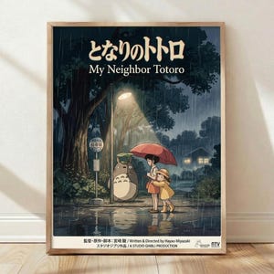 May include: Framed poster print of My Neighbor Totoro movie scene showing two children with umbrellas standing next to Totoro character at rainy bus stop under large tree, featuring Japanese text at top, dark blue and green tones with warm lighting, displayed in light wood frame leaning against white wall on wooden floor
