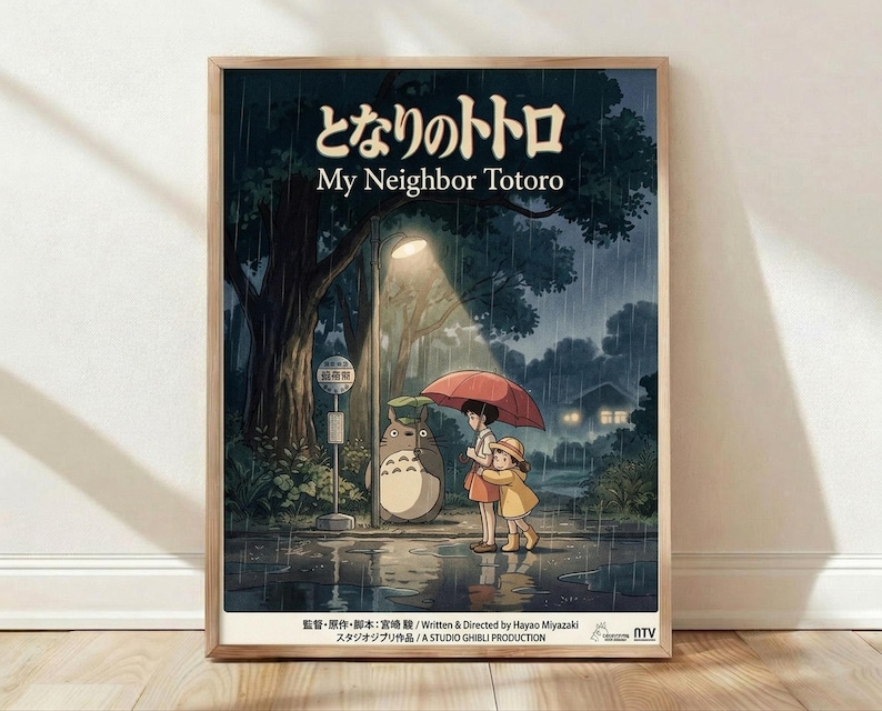 May include: Framed poster print of My Neighbor Totoro movie scene showing two children with umbrellas standing next to Totoro character at rainy bus stop under large tree, featuring Japanese text at top, dark blue and green tones with warm lighting, displayed in light wood frame leaning against white wall on wooden floor