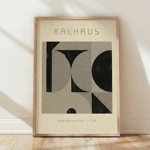 May include: A framed Bauhaus art print. The artwork features geometric shapes in black and gray on a beige background. The word "BAUHAUS" is printed at the top, and "Exhibition Art 1919" is printed at the bottom. The frame is made of light brown wood.