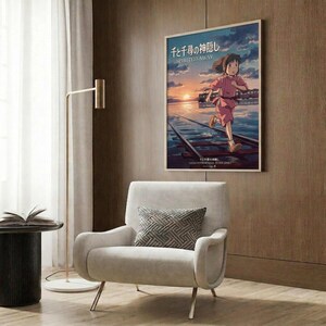 May include: Framed Spirited Away anime poster mounted on wood-paneled wall above cream upholstered armchair. Poster shows young girl in red outfit running across wooden bridge over water at sunset with blue and orange sky. Modern living room setting with gold floor lamp, geometric throw pillow, and dark side table with open book.