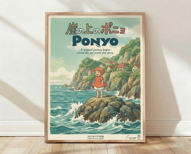 May include: Framed Studio Ghibli Ponyo poster in light wood frame, leaning against white wall on wooden floor. Artwork shows young girl with reddish hair in orange dress standing on rocky outcrop surrounded by ocean waves, with cliffside village houses visible in background. Japanese text at top, English subtitle reads "A magical journey begins where the sea meets the shore." Soft natural lighting creates gentle shadows on white textured wall.