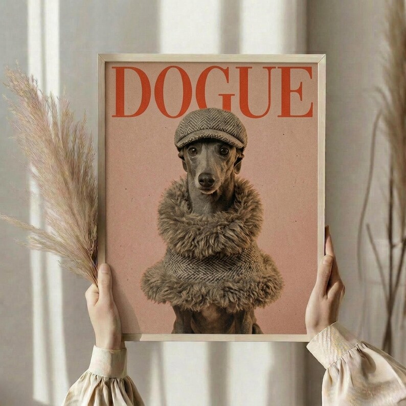 May include: Framed poster featuring a whippet dog wearing a tweed flat cap and fluffy fur collar against a peachy pink background, styled like a fashion magazine cover with "DOGUE" text in red letters. Person in cream ruffled sleeves holds the light wood-framed print, with dried wheat stems visible to the left against soft neutral curtains.