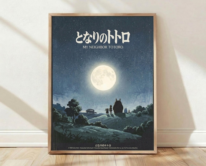 May include: Framed poster print leaning against white wall on wooden floor. Artwork shows nighttime scene with large full moon, starry blue sky, silhouettes of Totoro and children standing near Catbus on rolling hills with trees. Japanese text at top reads "My Neighbor Totoro" in white lettering. Natural wood frame, soft lighting with shadows cast on textured white wall.