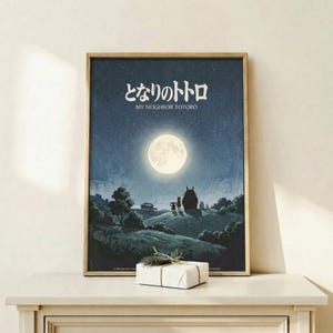 May include: Framed Studio Ghibli movie poster showing nighttime scene with full moon, starry sky, silhouettes of Totoro and children on rolling hills. Dark blue and teal tones with glowing white moon. Japanese text at top with English subtitle. Thin wooden frame displayed on light wood shelf against white wall, small wrapped gift package in front.