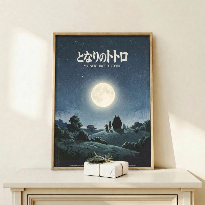 May include: Framed Studio Ghibli movie poster showing nighttime scene with full moon, starry sky, silhouettes of Totoro and children on rolling hills. Dark blue and teal tones with glowing white moon. Japanese text at top with English subtitle. Thin wooden frame displayed on light wood shelf against white wall, small wrapped gift package in front.