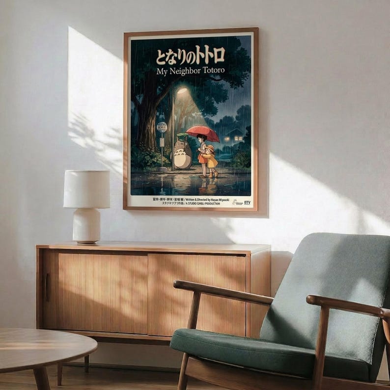 May include: Framed My Neighbor Totoro movie poster mounted on white wall above wooden sideboard with white table lamp. Poster shows Totoro and two children with red umbrella at rainy bus stop in forest. Mid-century modern living room with sage green armchair, round coffee table, and natural sunlight streaming across scene.
