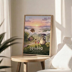 May include: Framed Studio Ghibli poster featuring Totoro character with umbrella standing beside two children in a whimsical landscape with rolling green hills, traditional Japanese house, floating airship, and sunset sky with pink and purple clouds. Wooden frame displayed on round wooden side table next to beige upholstered chair with plant shadows from window light creating soft ambiance on cream walls.