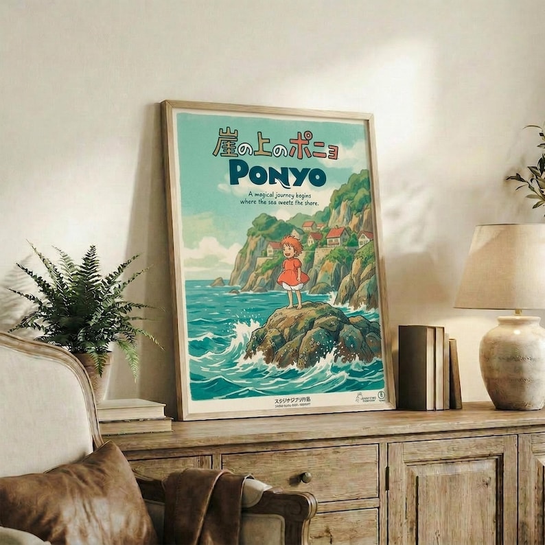 May include: Framed Studio Ghibli Ponyo poster displayed on rustic wooden credenza in living room setting. Poster features young girl in red dress standing on rocky outcrop surrounded by ocean waves, with Japanese text and coastal cliff scenery in soft blues and greens. Room includes fern plant, ceramic vase, books, and beige upholstered furniture with brown throw pillow.
