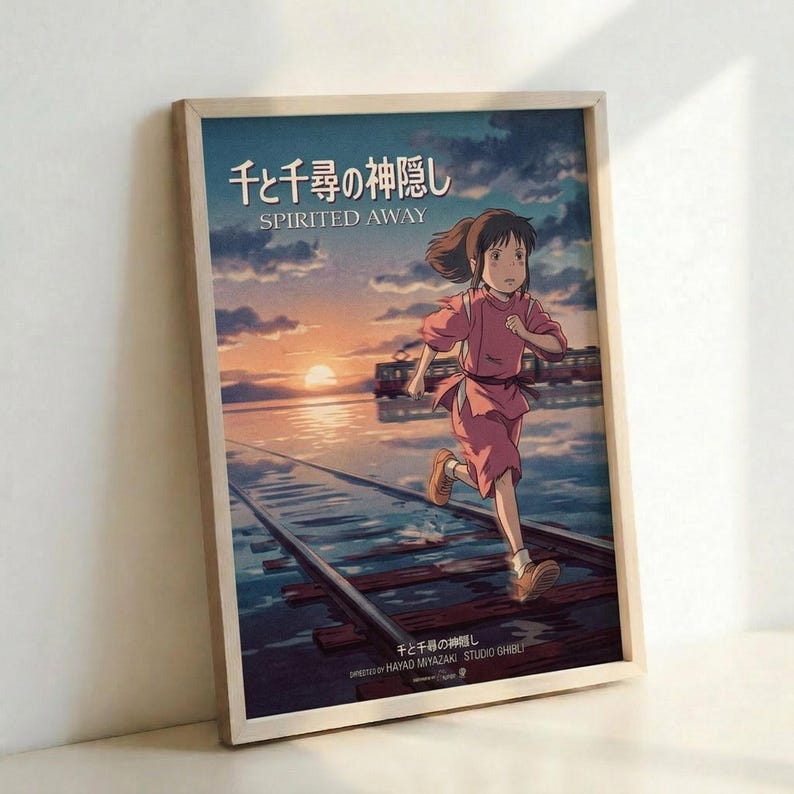 May include: Framed poster featuring animated character Chihiro from Studio Ghibli's Spirited Away running along flooded railway tracks at sunset. The artwork shows warm orange and pink sky colors reflecting on water, with Japanese text at top. Light wood frame displayed against white wall, leaning on surface with soft natural lighting creating subtle shadows.