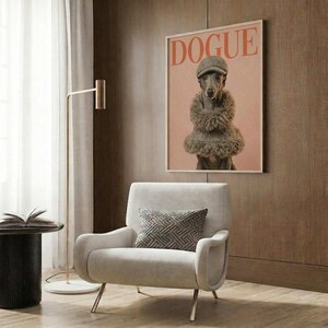 May include: Framed poster featuring a whippet dog wearing a textured cap and fur stole on pink background with "DOGUE" text in coral letters, displayed above a cream upholstered chair with geometric pillow in modern living room with wood paneling, brass floor lamp, and dark side table with open book.