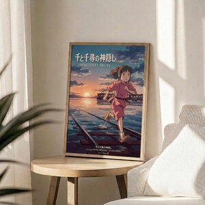 May include: Framed Spirited Away movie poster featuring animated girl with brown hair in pink outfit running on wooden railway tracks over water at sunset. Poster displays Japanese text and "SPIRITED AWAY" title with Studio Ghibli credits. Light wooden frame sits on round wooden side table next to beige upholstered chair and green plant leaves in bright, minimalist room setting.