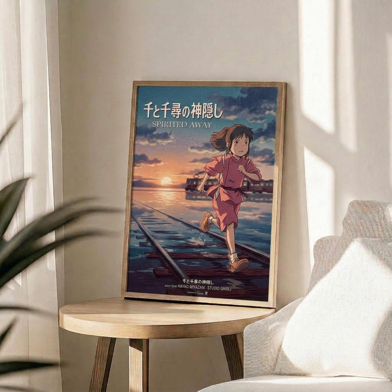 May include: Framed Spirited Away movie poster featuring animated girl with brown hair in pink outfit running on wooden railway tracks over water at sunset. Poster displays Japanese text and "SPIRITED AWAY" title with Studio Ghibli credits. Light wooden frame sits on round wooden side table next to beige upholstered chair and green plant leaves in bright, minimalist room setting.
