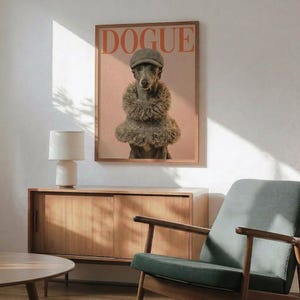 May include: A framed poster featuring a whippet dog wearing a grey flat cap and fluffy fur stole, styled as a fashion magazine cover with "DOGUE" text on peachy pink background. The wooden-framed print hangs on a white wall above a mid-century modern walnut credenza with white ceramic lamp. A mint green upholstered armchair with wooden frame sits nearby, with sunlight casting geometric shadows across the minimalist room setting.