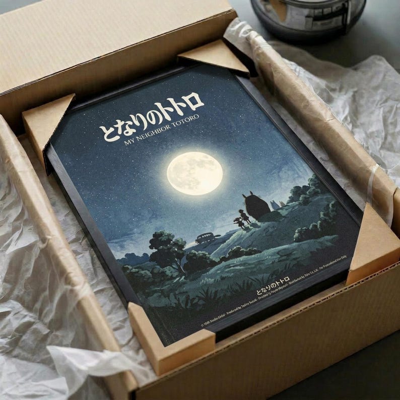 May include: Framed poster showing moonlit nighttime scene with full moon, starry sky, and silhouetted landscape featuring trees and small figures. Japanese text at top reads "My Neighbor Totoro." Art print nestled in cardboard shipping box with protective tissue paper wrapping. Dark blue and teal color palette with cream moon creating atmospheric lighting across rolling hills and vegetation.
