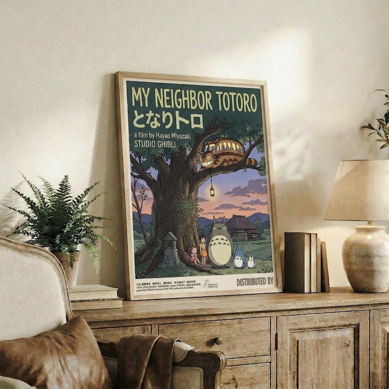 May include: Framed vintage-style Studio Ghibli poster displayed on rustic wooden sideboard in cozy living room setting. Poster shows Totoro characters under large tree with Catbus in branches, rendered in muted earth tones against twilight sky. Scene includes fern plant, ceramic vase, stacked books, and cream-colored armchair with brown throw pillows.