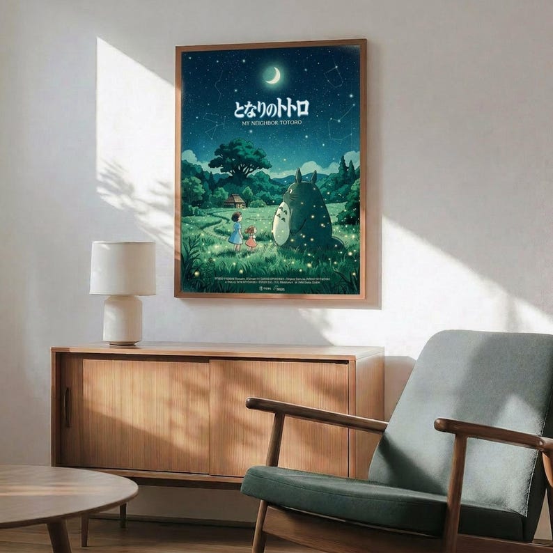 May include: Framed My Neighbor Totoro poster mounted on white wall above wooden credenza. Artwork shows nighttime scene with starry sky, crescent moon, children and large Totoro character in grassy field. Mid-century modern room setting with sage green armchair, round coffee table, and table lamp. Natural sunlight creates shadows across the space. Poster has thin brown frame.