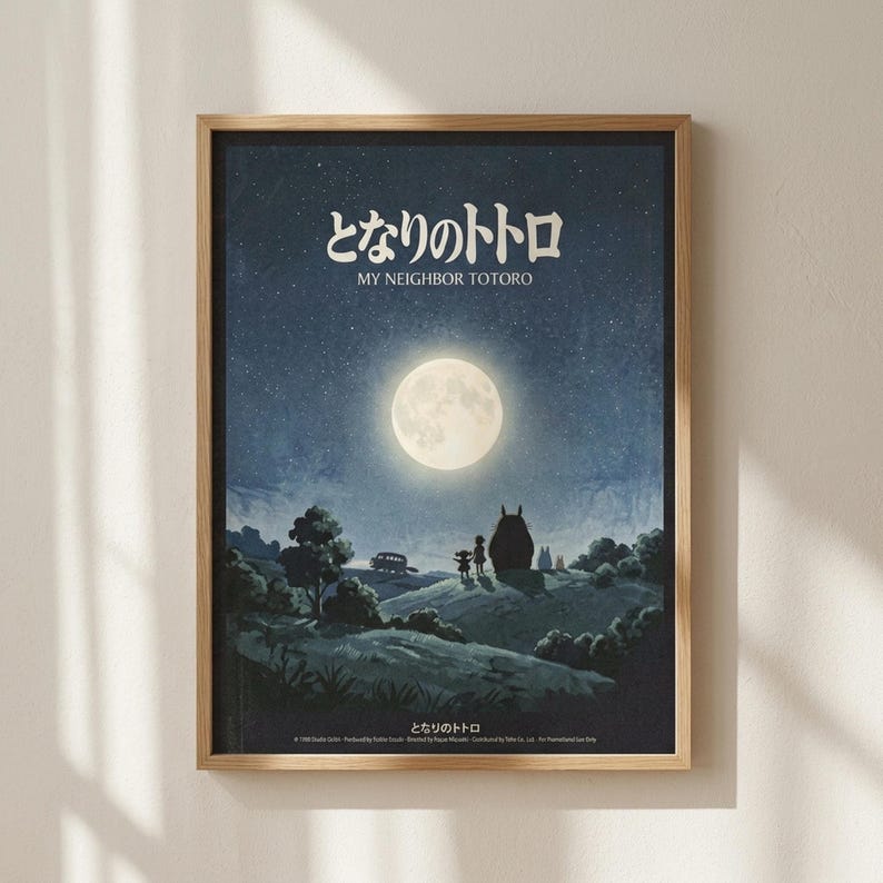 May include: Framed poster mounted on white wall with natural lighting creating shadows. The artwork features a nighttime scene with a large full moon in a starry blue sky. Silhouettes of two children and the iconic large Totoro character stand on a hill surrounded by trees and countryside landscape. Japanese text appears at the top with English translation below. The poster has a thin light wood frame and displays rich blues and greens with the bright white moon as focal point.