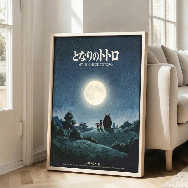 May include: Framed poster displaying a moonlit night scene with two silhouetted children standing beside a large creature on a hill, featuring Japanese text at top and English subtitle below. Dark blue starry sky with bright full moon illuminates rolling countryside landscape. Light wood frame leans against white wall beside beige upholstered chair in bright room with large windows.
