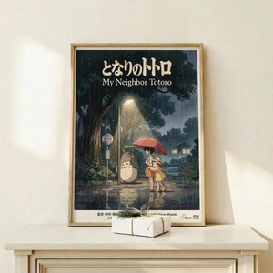 May include: Framed poster of Studio Ghibli's My Neighbor Totoro showing two children with red umbrella standing at rainy bus stop beside large gray Totoro character. Dark moody forest scene with glowing lights in background. Natural wood frame displayed on light wooden shelf with small wrapped gift box. Warm beige wall behind.