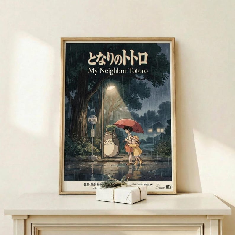 May include: Framed poster of Studio Ghibli's My Neighbor Totoro showing two children with red umbrella standing at rainy bus stop beside large gray Totoro character. Dark moody forest scene with glowing lights in background. Natural wood frame displayed on light wooden shelf with small wrapped gift box. Warm beige wall behind.