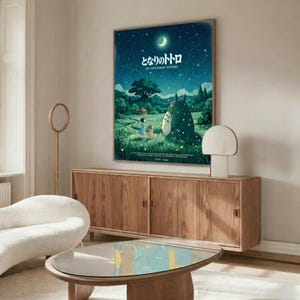 May include: Studio Ghibli My Neighbor Totoro movie poster displayed on white wall above wooden sideboard in modern living room. Poster features nighttime forest scene with children, large gray Totoro character, crescent moon and stars in deep blue-green tones. Room includes cream upholstered chair, glass-top coffee table, and minimalist decor with natural wood finishes throughout.