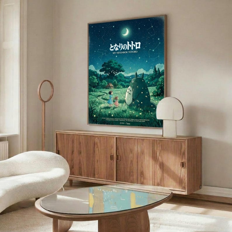 May include: Studio Ghibli My Neighbor Totoro movie poster displayed on white wall above wooden sideboard in modern living room. Poster features nighttime forest scene with children, large gray Totoro character, crescent moon and stars in deep blue-green tones. Room includes cream upholstered chair, glass-top coffee table, and minimalist decor with natural wood finishes throughout.