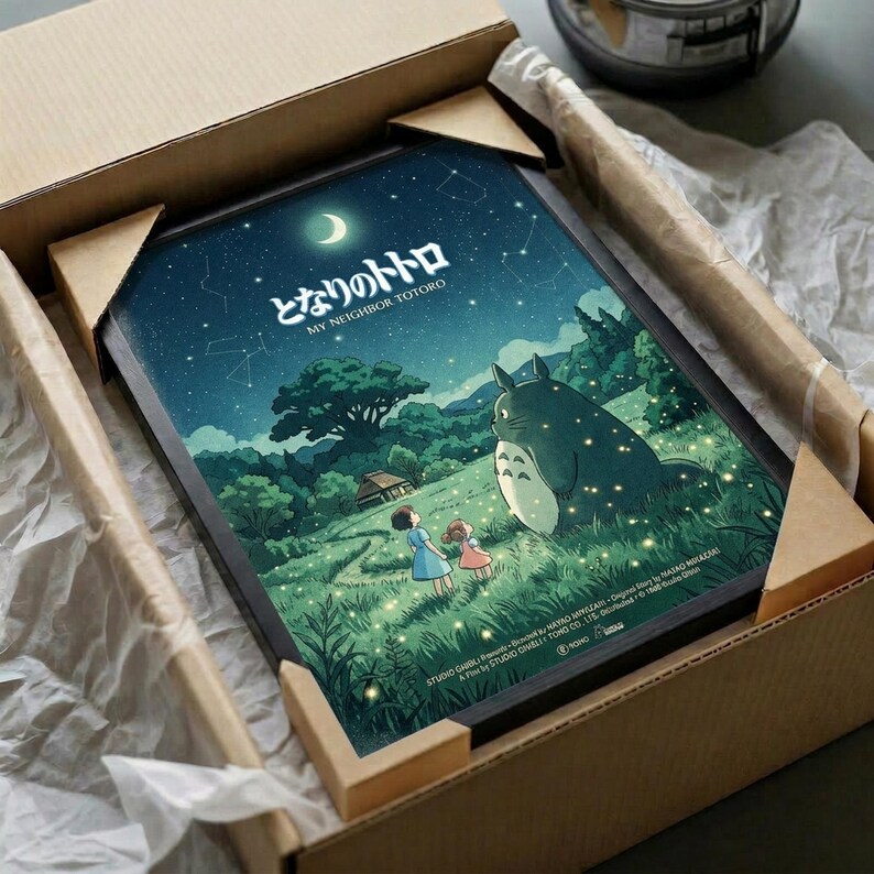 May include: Framed poster print in cardboard box with protective wrapping. Artwork shows nighttime scene with two children standing near large gray forest creature under starry sky with crescent moon. Green hills, trees, and small house visible in background. Warm yellow lights scattered throughout scene. Japanese text at top. Print appears to be in black frame nestled in brown shipping box with white tissue paper padding.