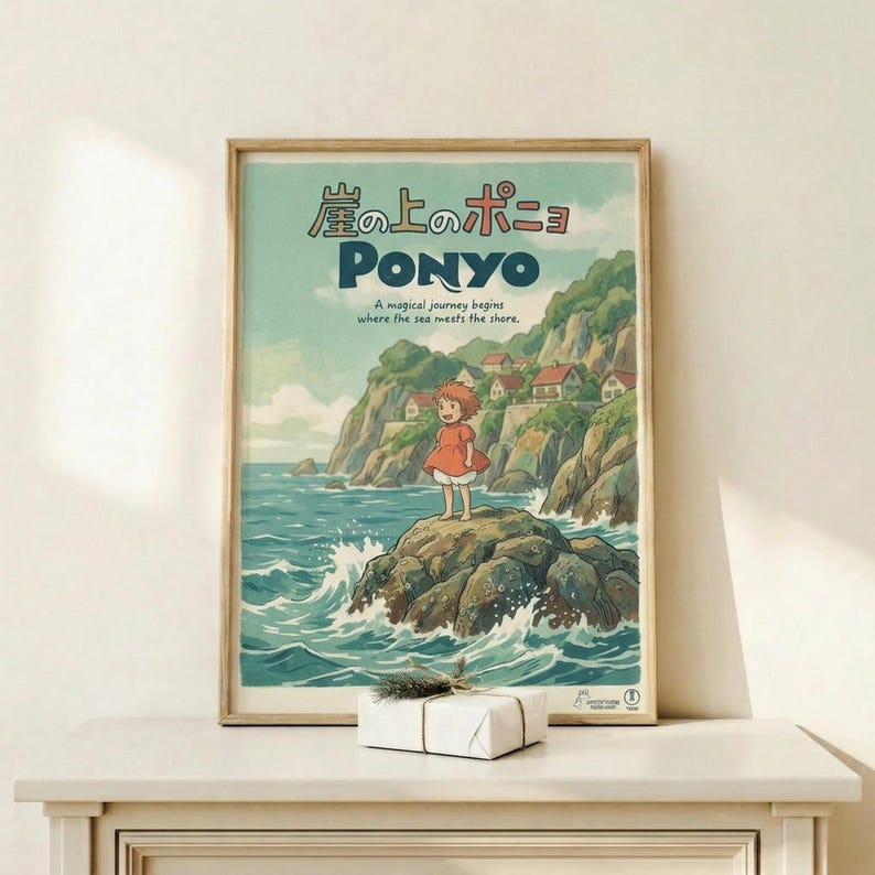 May include: Framed Studio Ghibli Ponyo poster displayed on light wood shelf against white wall. The colorful illustrated print shows a red-haired girl in orange dress standing on rocky coastline with crashing waves, clifftop village in background. Japanese and English text at top reads "A magical journey begins where the sea meets the shore." Natural wood frame, small white gift box with twine sits in front. Soft natural lighting creates gentle shadows on minimalist display.