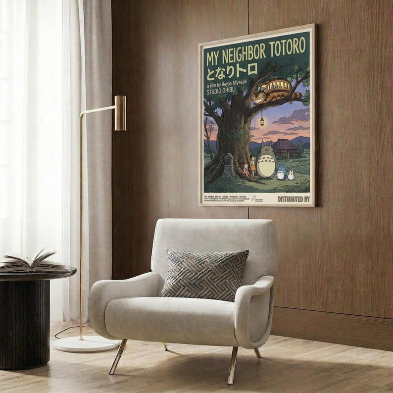 May include: Framed My Neighbor Totoro movie poster displayed on wood-paneled wall above modern beige armchair with geometric pillow. Poster shows iconic characters under large tree at twilight with Catbus in branches. Contemporary living room setting with brass floor lamp and side table.