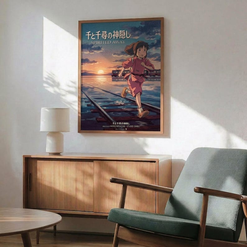 May include: Framed Spirited Away movie poster mounted on white wall above wooden sideboard with table lamp. Shows animated girl character running on wooden planks over water at sunset with Japanese text and "Spirited Away" title. Natural wood frame matches mid-century modern furniture including sage green upholstered armchair. Warm sunlight creates shadows across minimalist living room setting.