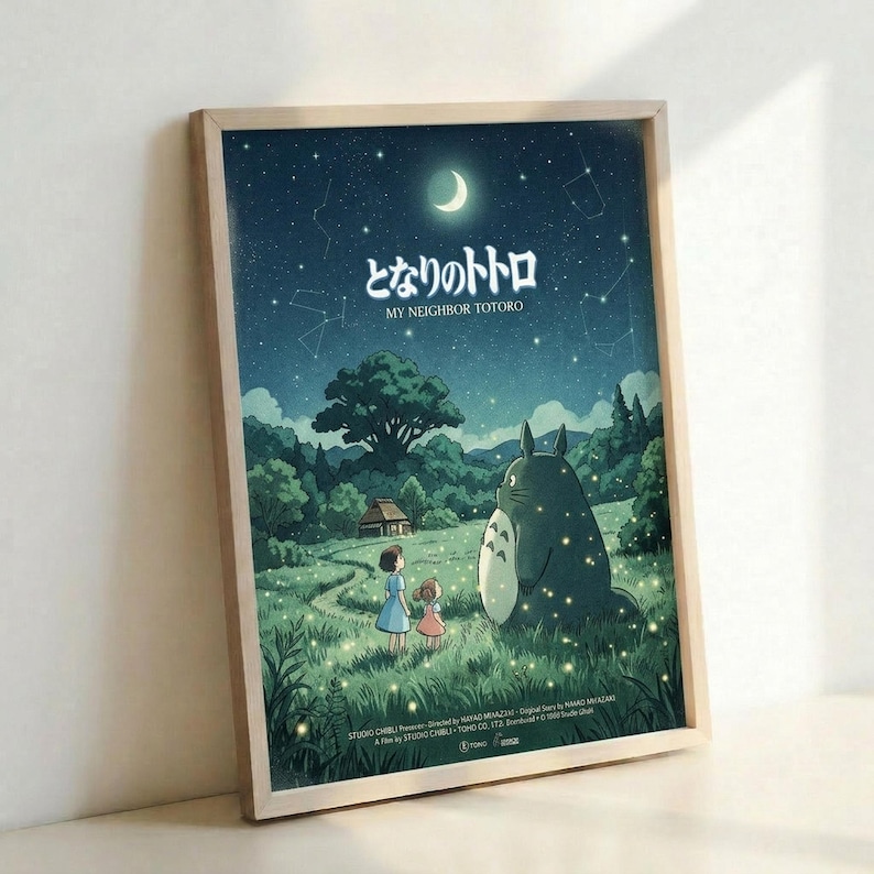 May include: Framed poster print leaning against white wall showing nighttime scene with two children standing beside large gray Totoro character in grassy field. Starry sky with crescent moon and constellations above, green trees and small house in background. Japanese text at top reads "My Neighbor Totoro" with Studio Ghibli credits below. Light wood frame, fireflies scattered throughout scene.