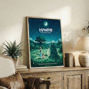 May include: Framed Studio Ghibli My Neighbor Totoro poster displayed on rustic wooden sideboard in cozy living room setting. Poster features nighttime scene with starry sky, crescent moon, two children and large Totoro character in green meadow landscape. Natural lighting illuminates the space with fern plant, books, ceramic vase, and beige armchair with brown leather cushion visible in warm, neutral-toned room.