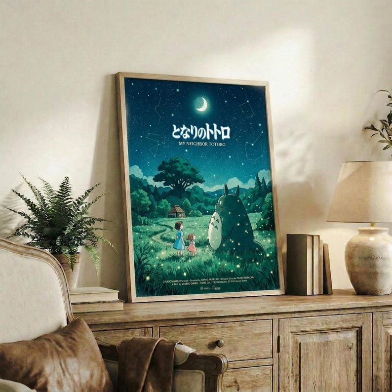May include: Framed Studio Ghibli My Neighbor Totoro poster displayed on rustic wooden sideboard in cozy living room setting. Poster features nighttime scene with starry sky, crescent moon, two children and large Totoro character in green meadow landscape. Natural lighting illuminates the space with fern plant, books, ceramic vase, and beige armchair with brown leather cushion visible in warm, neutral-toned room.